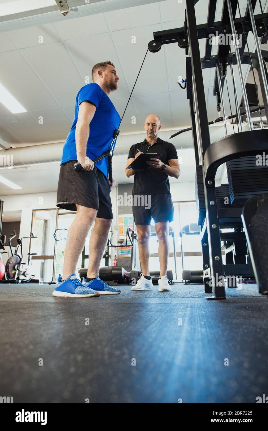A low-down shot of a mid-adult caucasian man training in the gym, he is strengthening his triceps. A fitness instructor can be seen standing by the ma Stock Photo