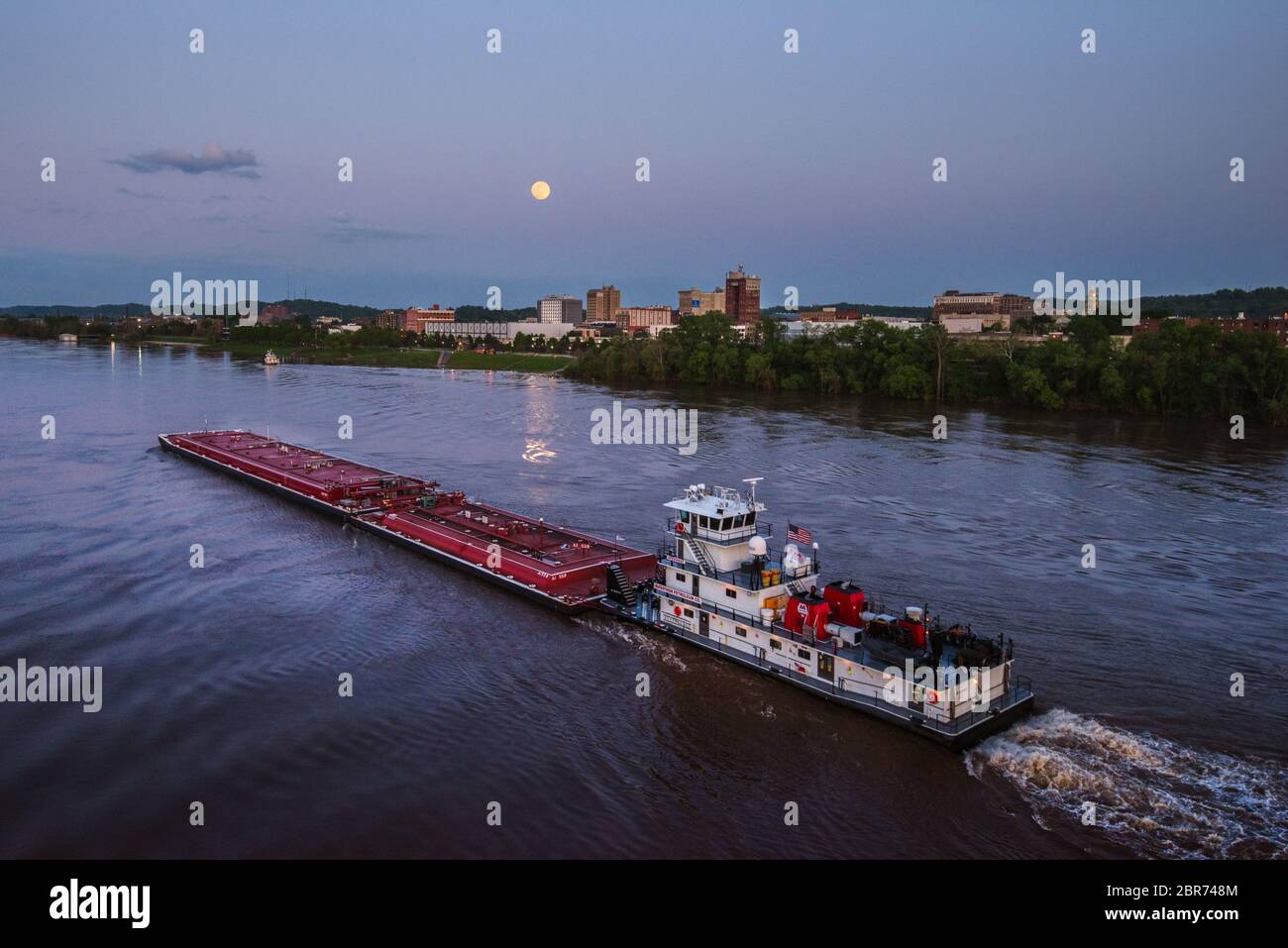 The moon rises above the river front park in Huntington, West Virginia with a large boat passing through the Ohio River. Stock Photo