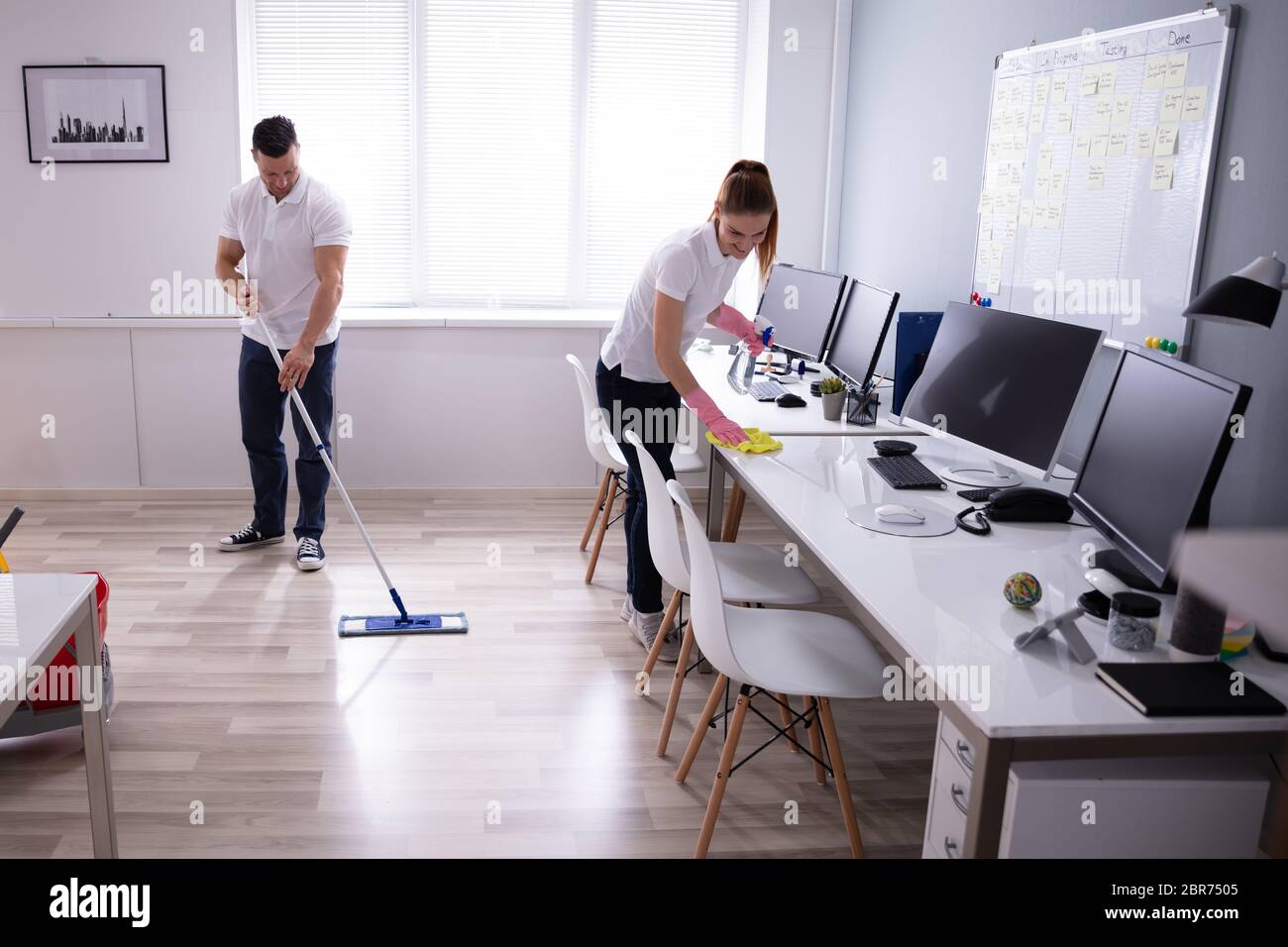 Two Smiling Young Janitor Cleaning The Desk And Mopping Floor In The Office Stock Photo