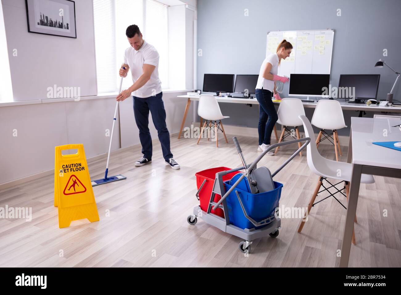 Two Smiling Young Janitor Cleaning The Desk And Mopping Floor In The Office Stock Photo