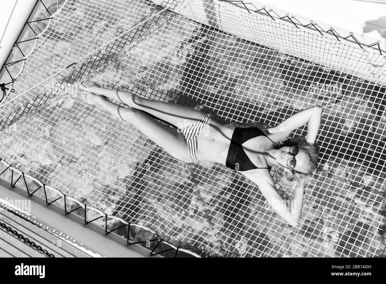 Womanin bikini tanning and relaxing on a summer sailin cruise, lying in hammock of luxury catamaran boat. Black and white image. Stock Photo