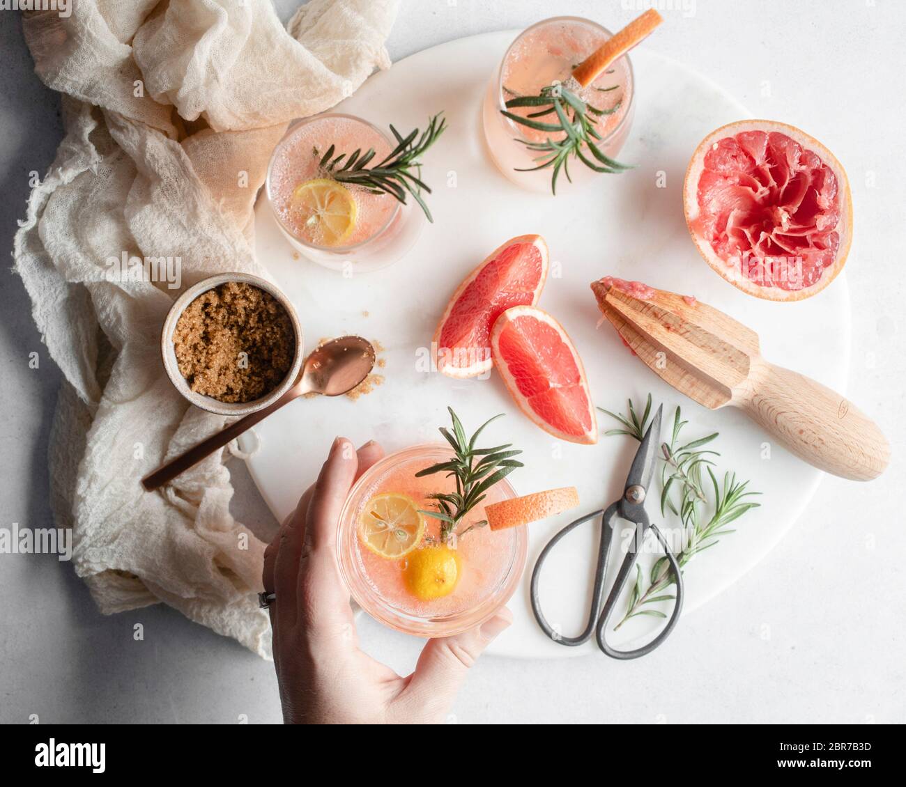 Overhead shot a hand reaching for a glass of grapefruit cocktail on a round platter with rosemary and grapefruit slices and a cup of brown sugar with Stock Photo
