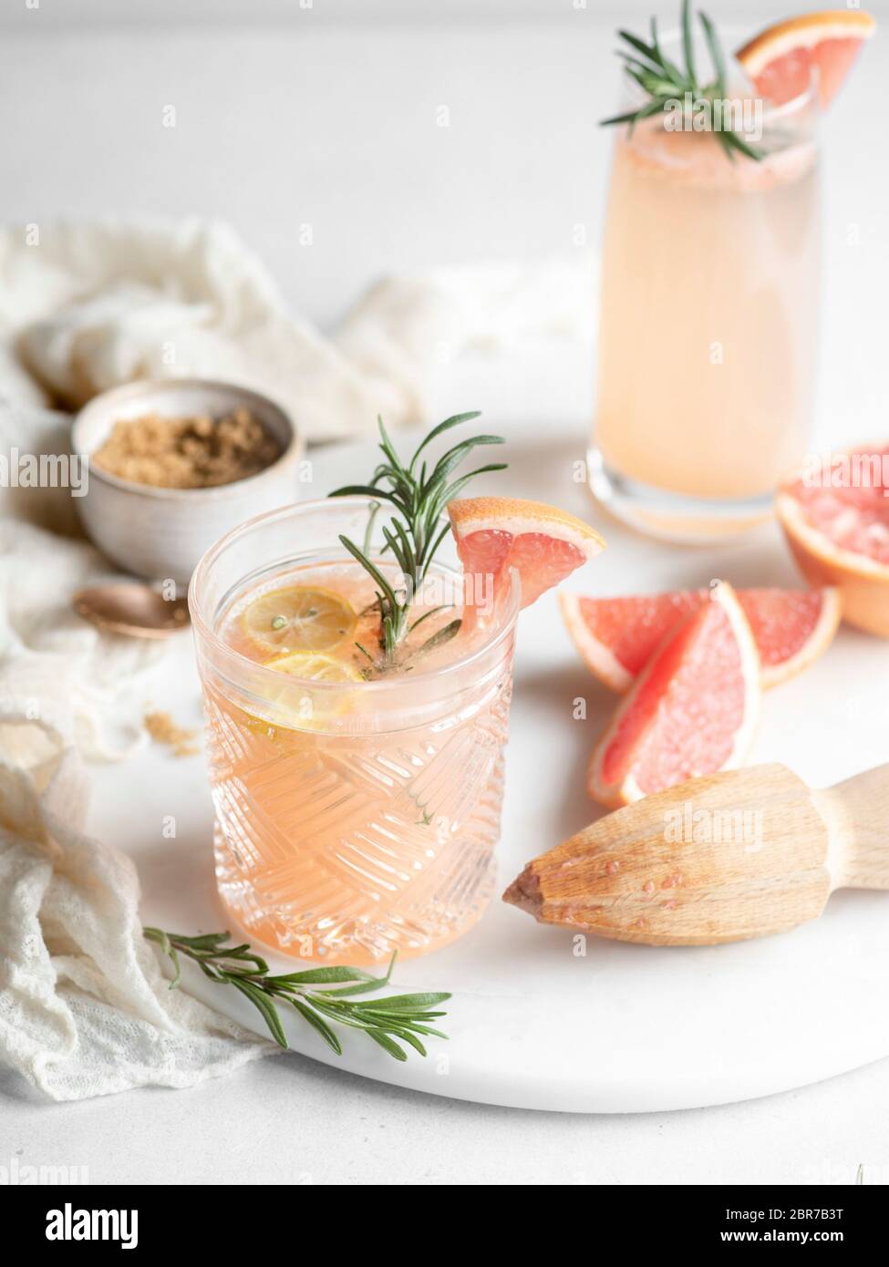 Old fashioned drinking glass filled with a grapefruit juice cocktail and garnished with lemon and rosemary on a white tray with a wooden hand juicer Stock Photo