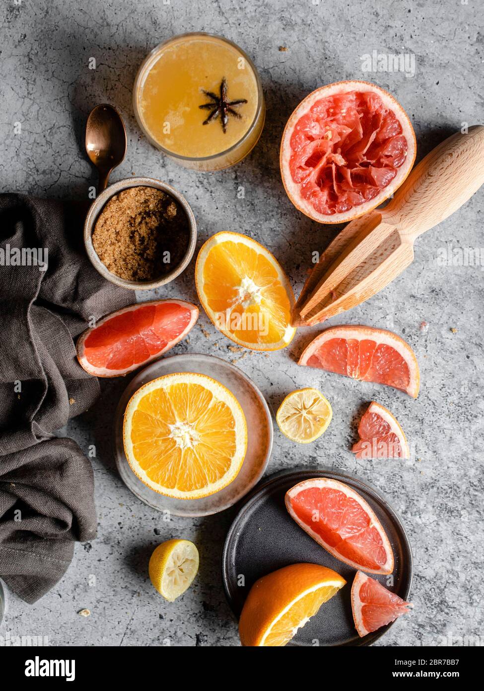 Overhead shot of sliced and halved oranges and grapefruits on a grey marble countertop, surrounded by a wooden hand juicer, a pocket knife, a grey nap Stock Photo