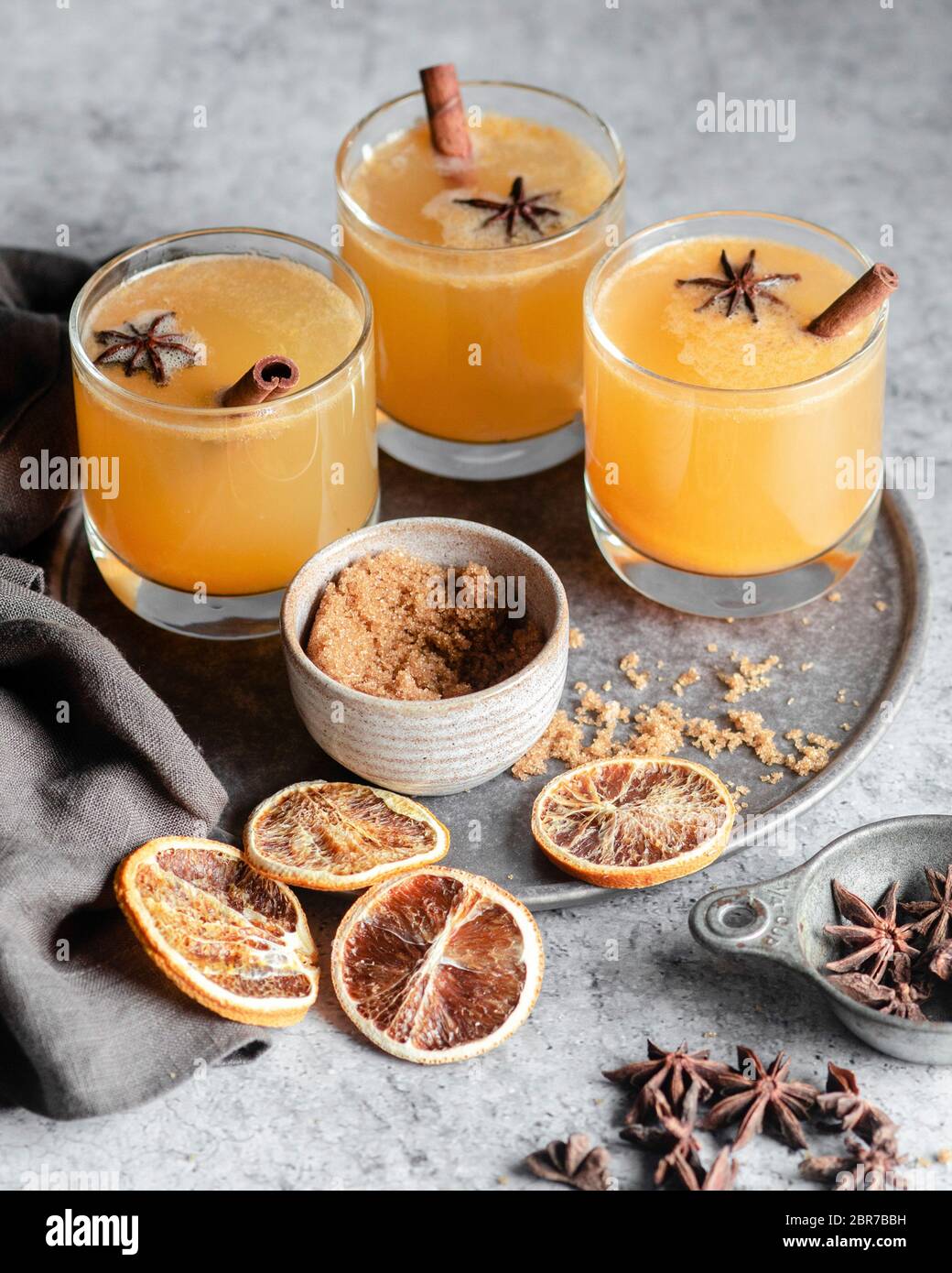 Three glasses of an orange spiced drink on a silver tray, garnished with star anise and cinnamon sticks and surrounded by blood oranges, brown sugar, Stock Photo