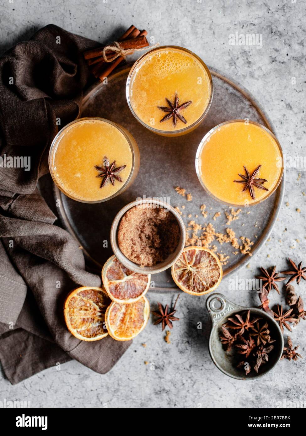 Overhead shot of three glasses of an orange spiced drink on a silver tray, garnished with star anise and surrounded by blood oranges, cinnamon sticks, Stock Photo