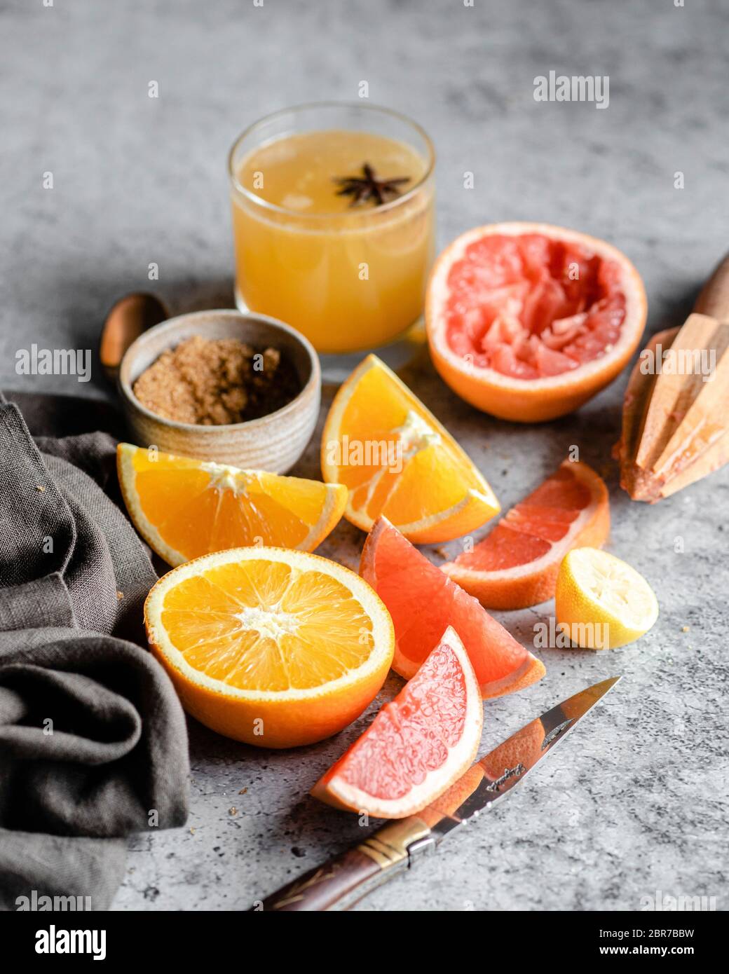 Sliced and halved oranges and grapefruits on a grey marble countertop, surrounded by a wooden hand juicer, a pocket knife, a grey napkin, a bowl of br Stock Photo