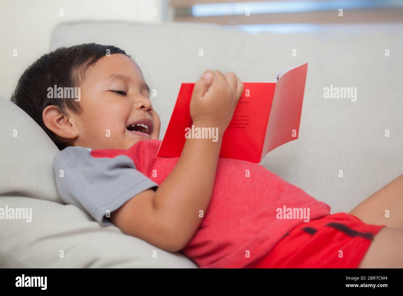 A young reader holding a simple book in his hand and reading out loud while relaxed on a couch. Stock Photo