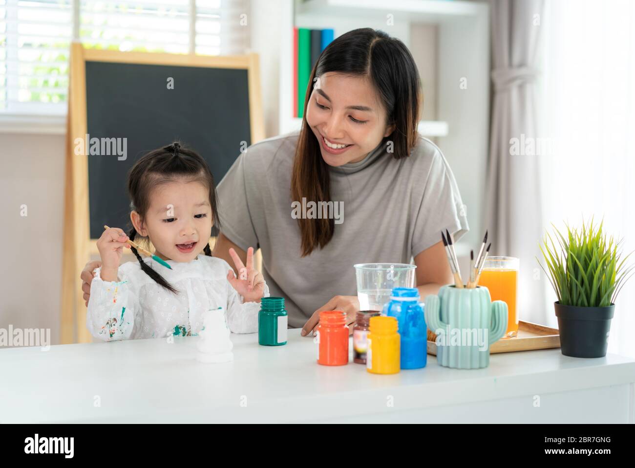 Asian kindergarten school girl with mother painting Plaster doll with Acrylic water color paint in living room at home. Homeschooling and distance lea Stock Photo
