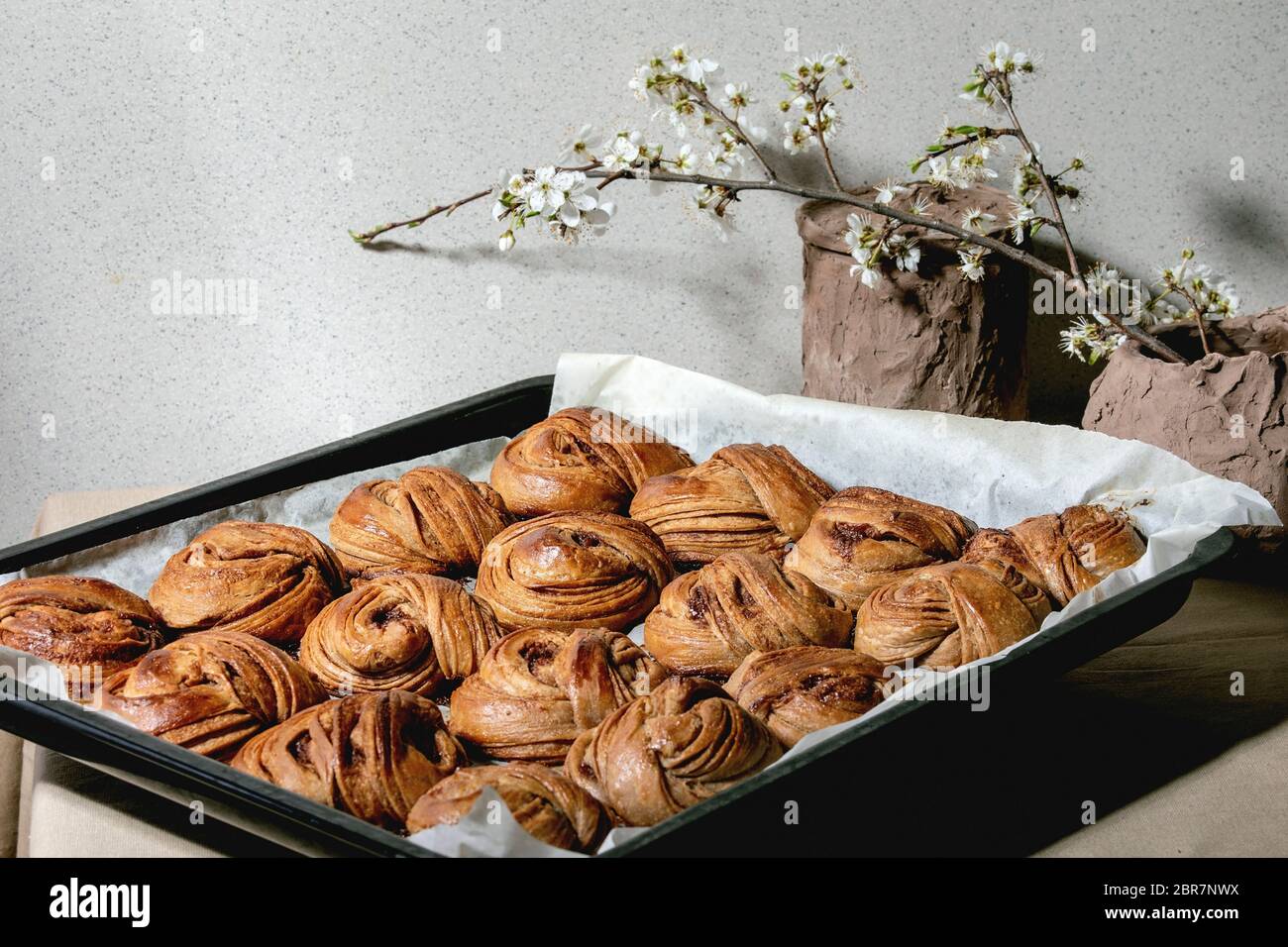 Fresh baked traditional Swedish cinnamon sweet buns Kanelbulle on oven tray cover by baking paper on beige linen table cloth. Blossom branches behind Stock Photo