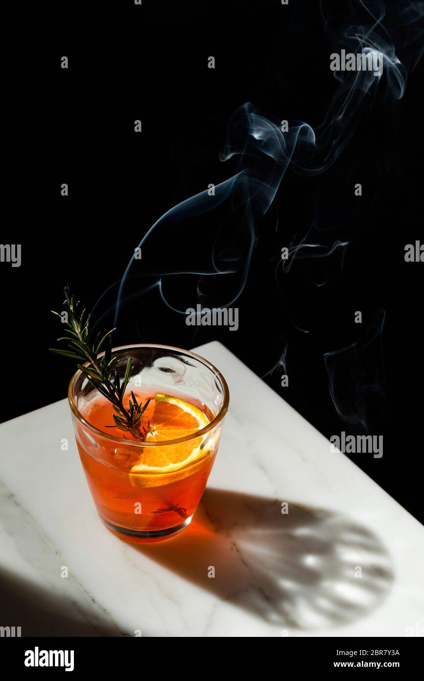 Negroni cocktail with a slice of orange and a smoking rosemary sprig garnish. The view features the corner of a marble tabletop with the shadow of the Stock Photo