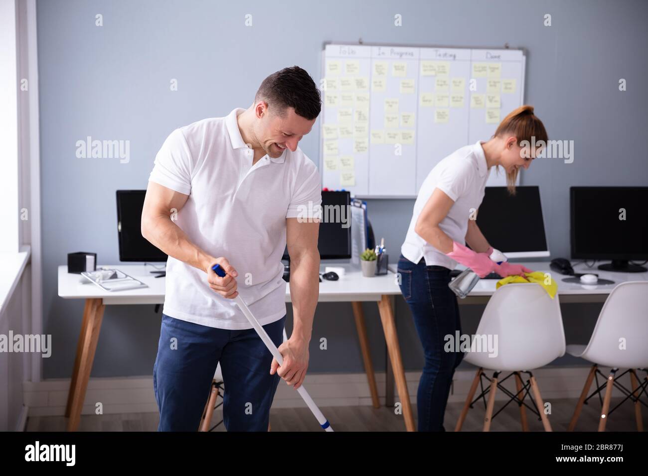 Two Smiling Young Janitor Cleaning The Desk And Mopping Floor In The Office Stock Photo