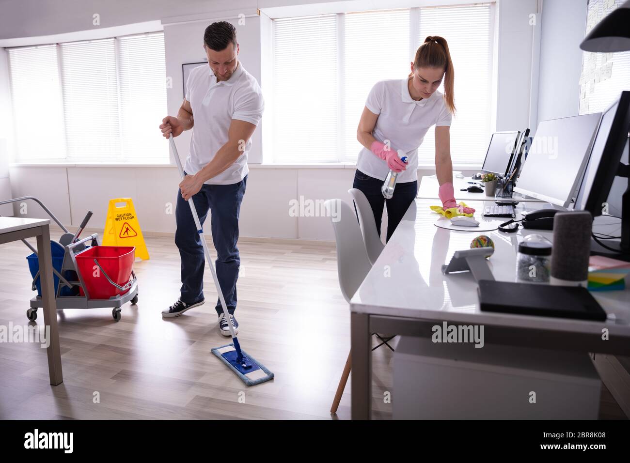 Two Smiling Young Janitor Cleaning The Desk And Mopping Floor In The Office Stock Photo