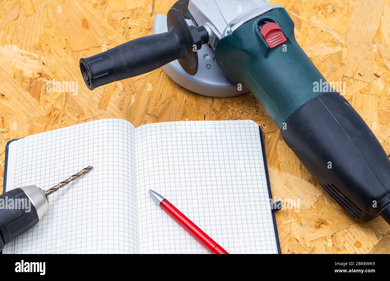 The image shows an angle grinder with a driller and notebook on a wooden table Stock Photo
