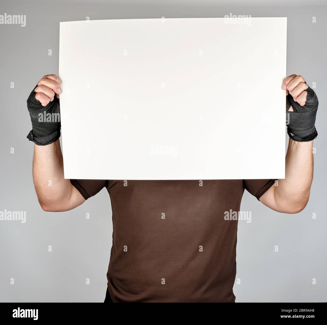 athlete in brown clothes holds a large blank white sheet of paper, his hands are wrapped in a black sports textile bandage, copy space Stock Photo