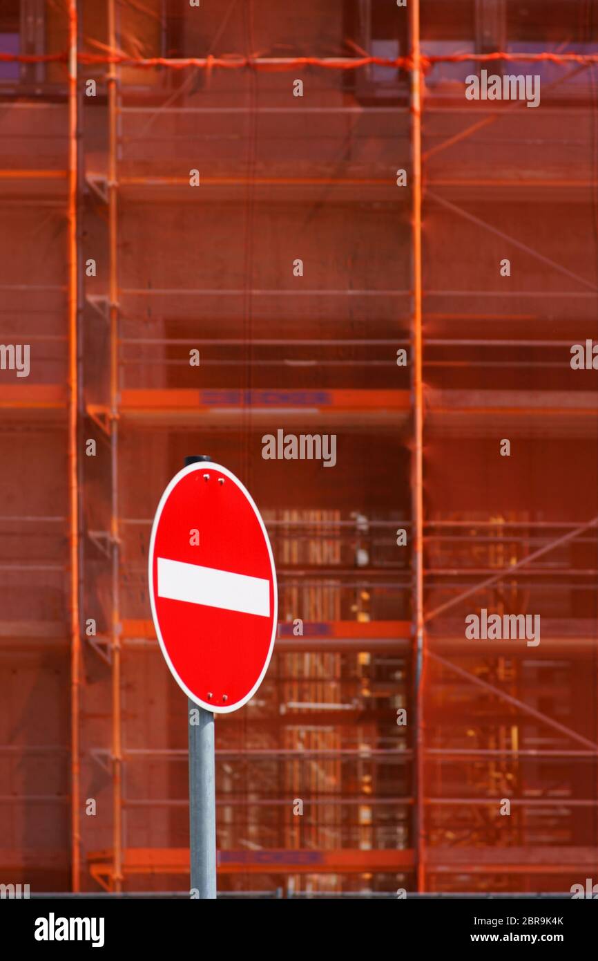 The close-up of a scaffolding with buttresses and safety nets in front of which stands a red one-way street sign. Stock Photo