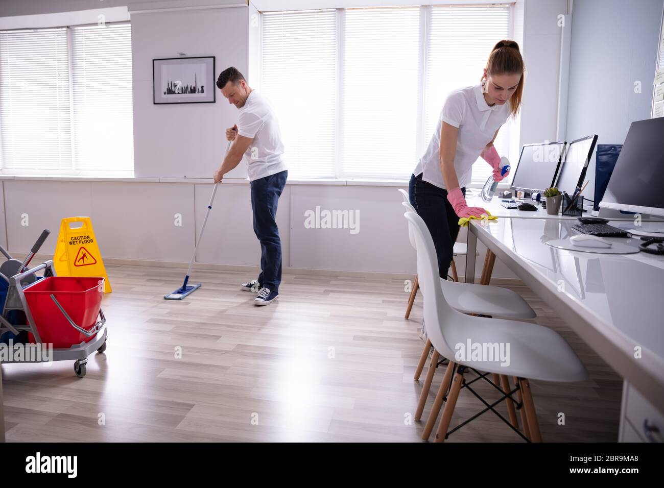 Two Smiling Young Janitor Cleaning The Desk And Mopping Floor In The Office Stock Photo