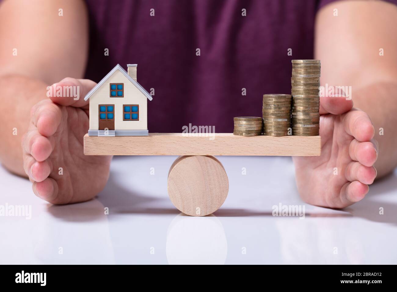 A Person's Hand Protecting Balance Between House Model And Money Coins On A Wooden Seesaw Stock Photo