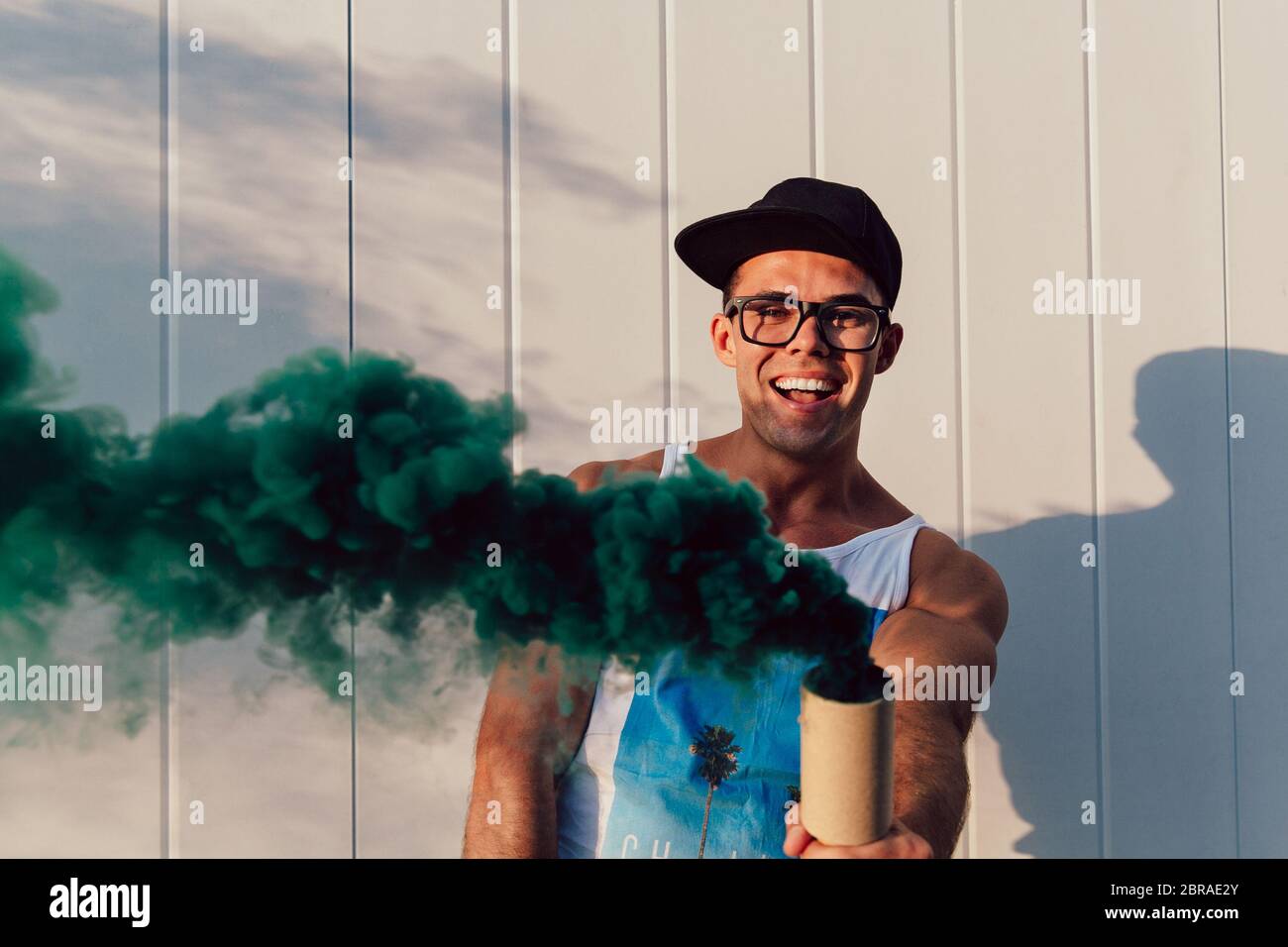 Cheerful attractive young man standing with green smoke flare outdoors, wearing trendy clothes, eyeglasses and rap cap. Stock Photo