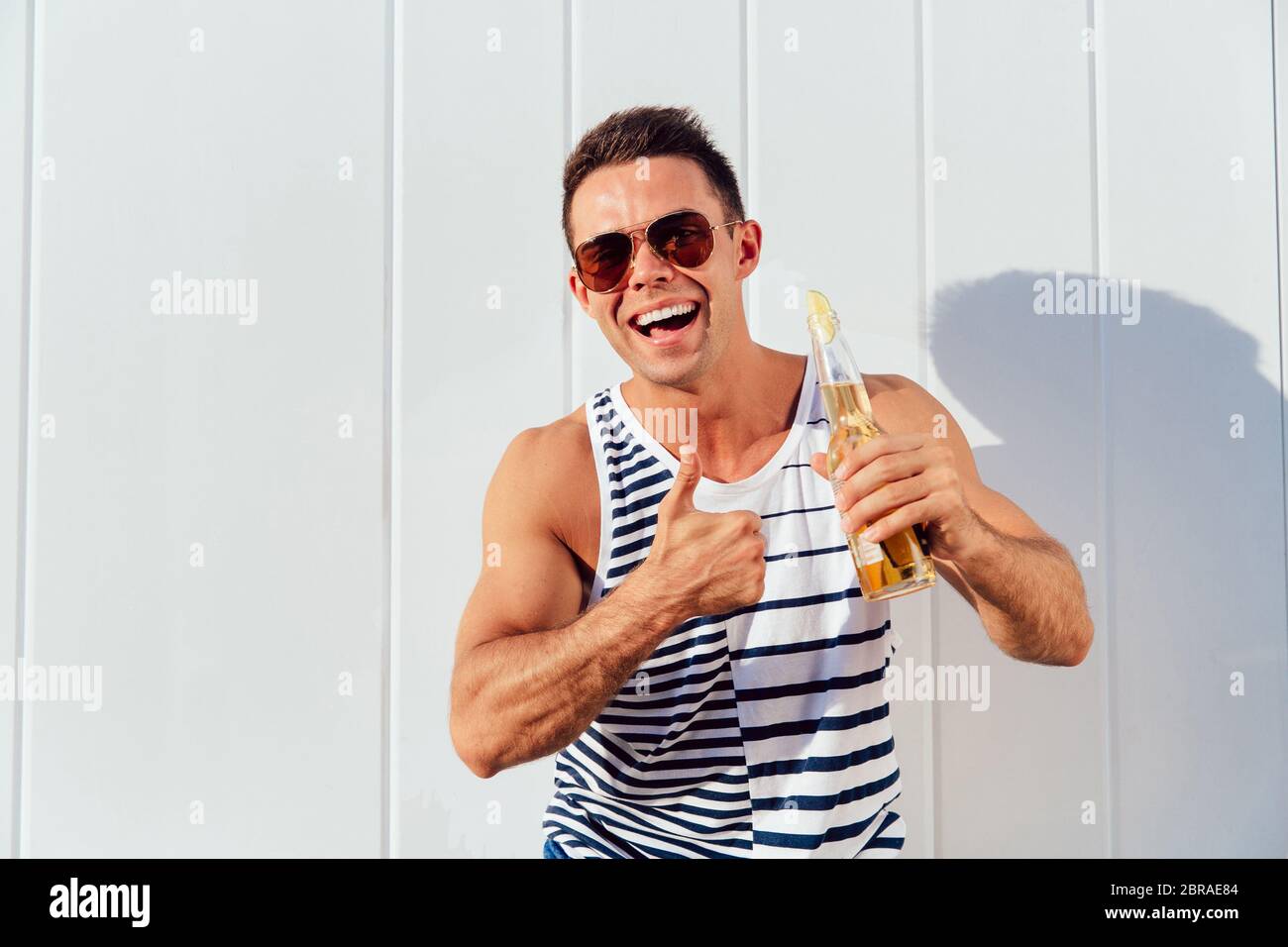 Happy young man in sunglasses showing a thumb up, while standing with bottle of cold beer, outdoors. Dressed in stylish clothes. Stock Photo
