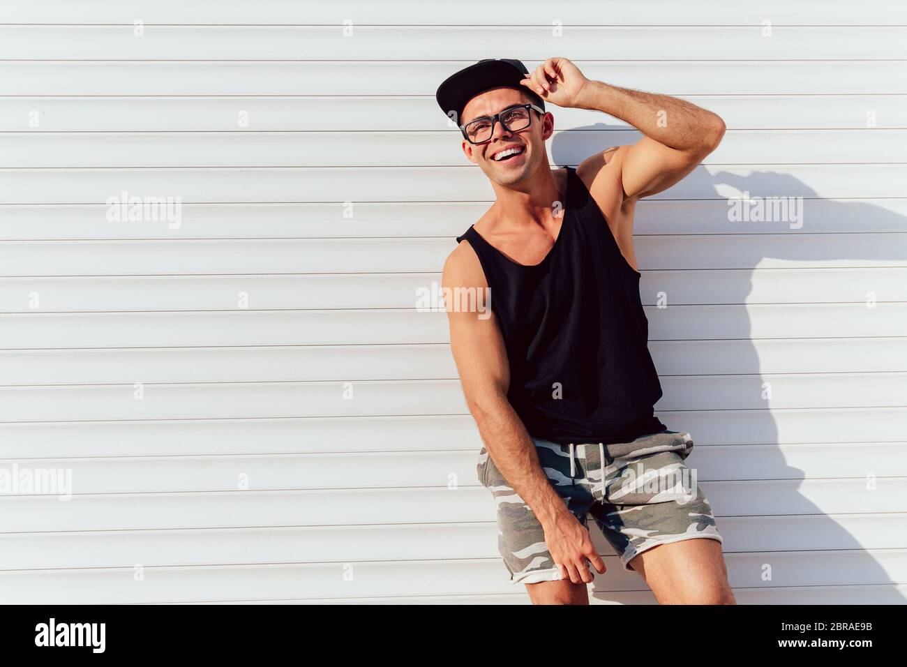 Funny attractive man in eyeglasses posing near the urban wall, wearing black singlet, shorts and cap. Outdoors. Stock Photo