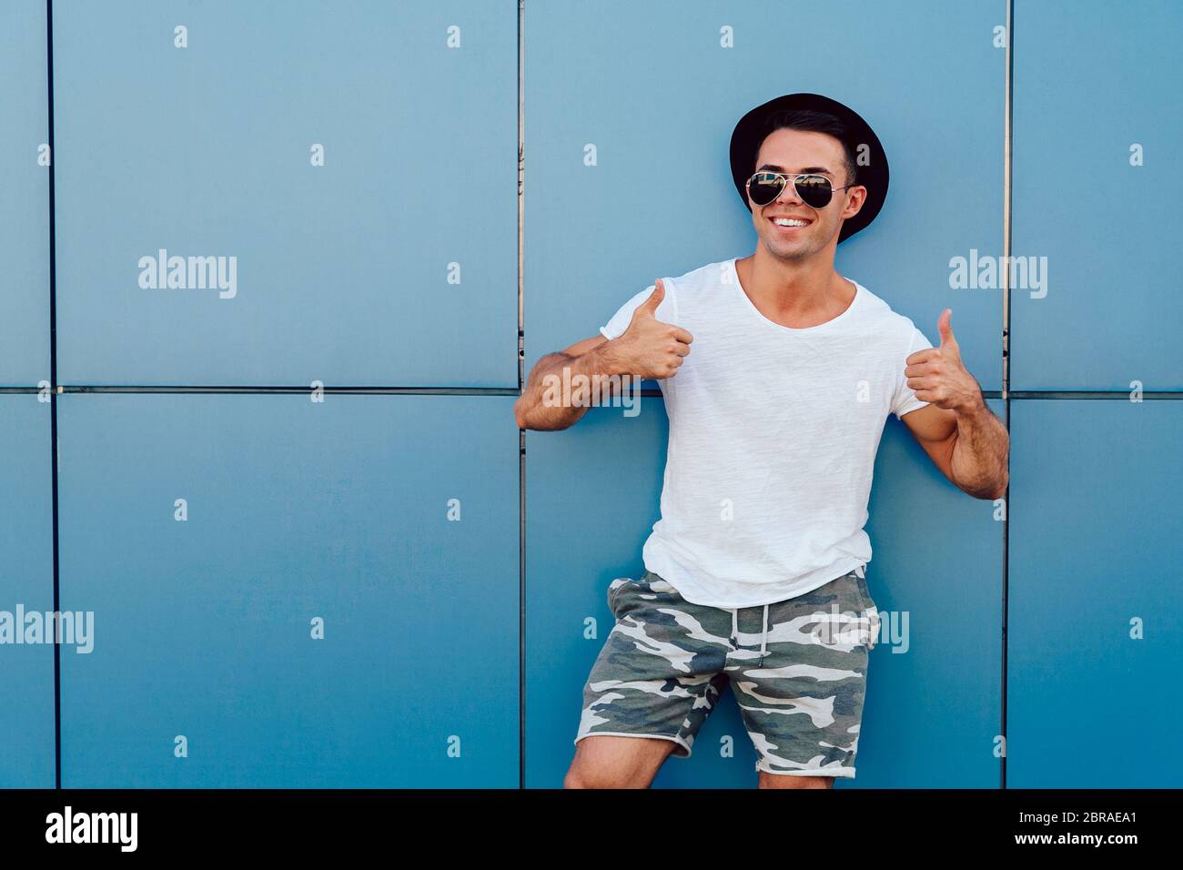 Happy, handsome man in sunglasses showing thumb up on two hands, standing against the wall, outdoors. Dressed in stylish clothes and hat. Stock Photo