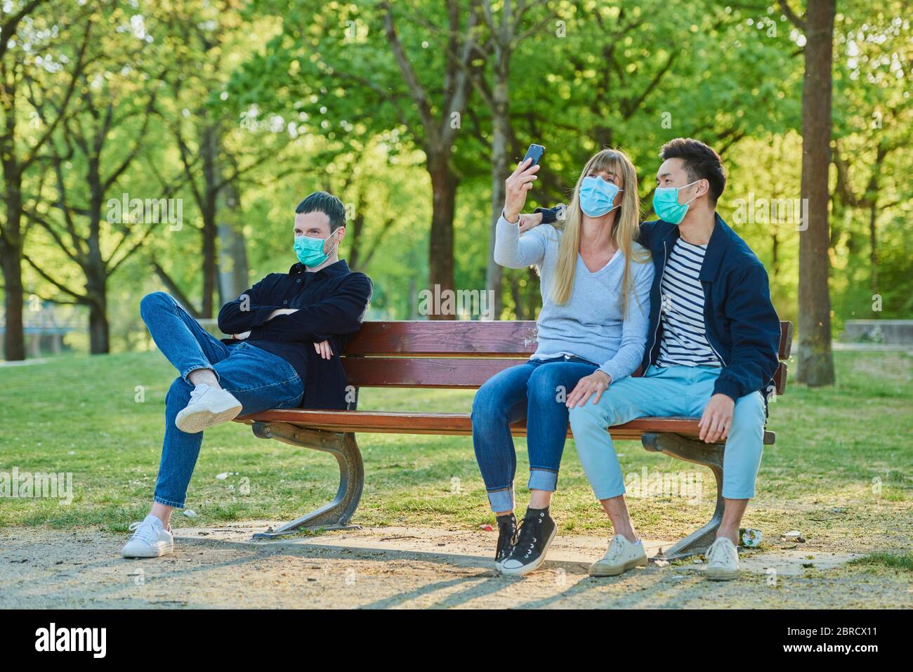 Ein Mann und ein Paar mit Mundschutzmaske auf einer Parkbank in einem Stadtpark sitzend, Corona-Krise, Regensburg, Bayern, Deutschland Stock Photo