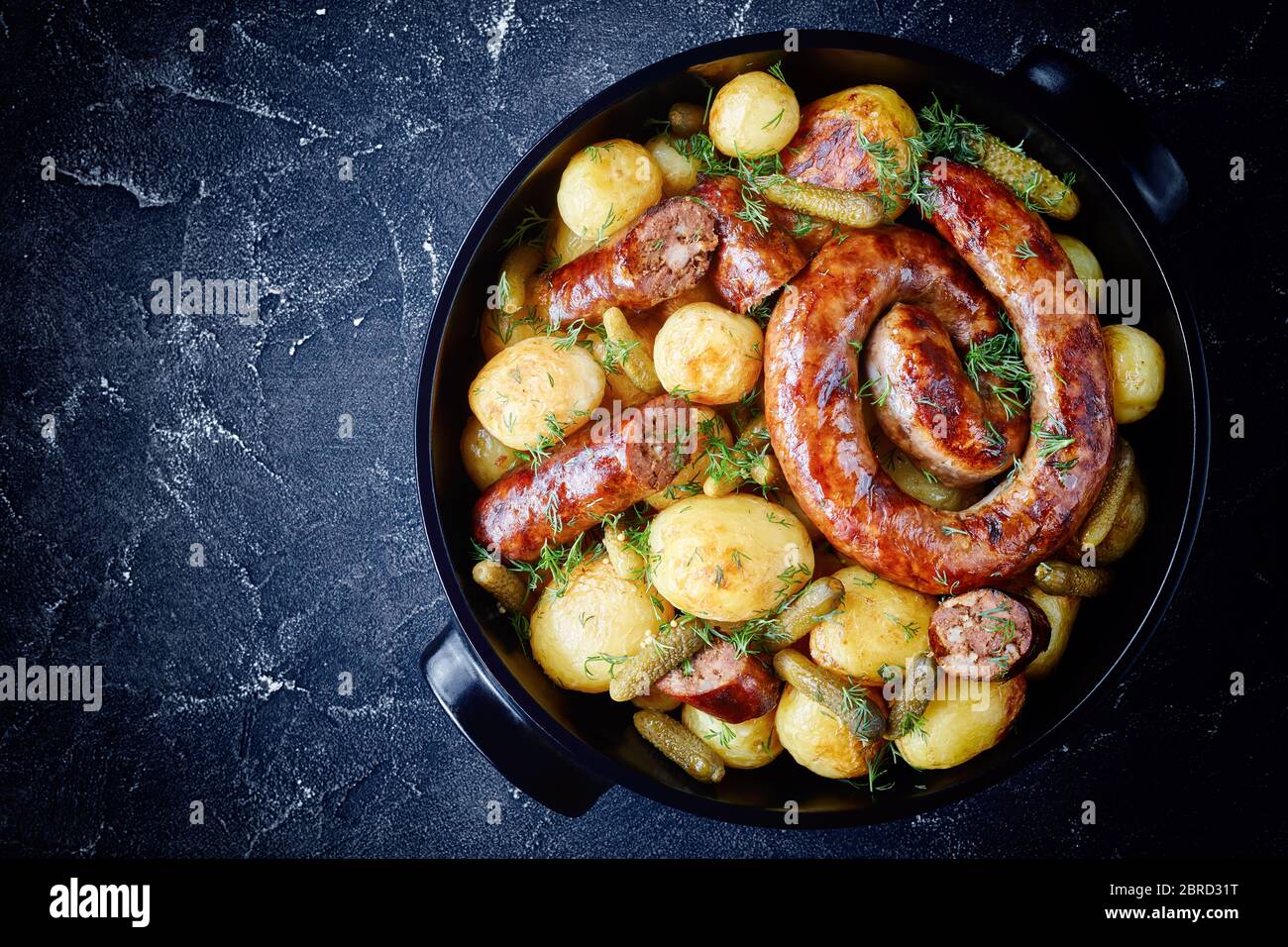 Black baking dish with new potato, round italian pork sausage with gherkins, sprinkled with fresh dill on a dark concrete background, top view, flat l Stock Photo