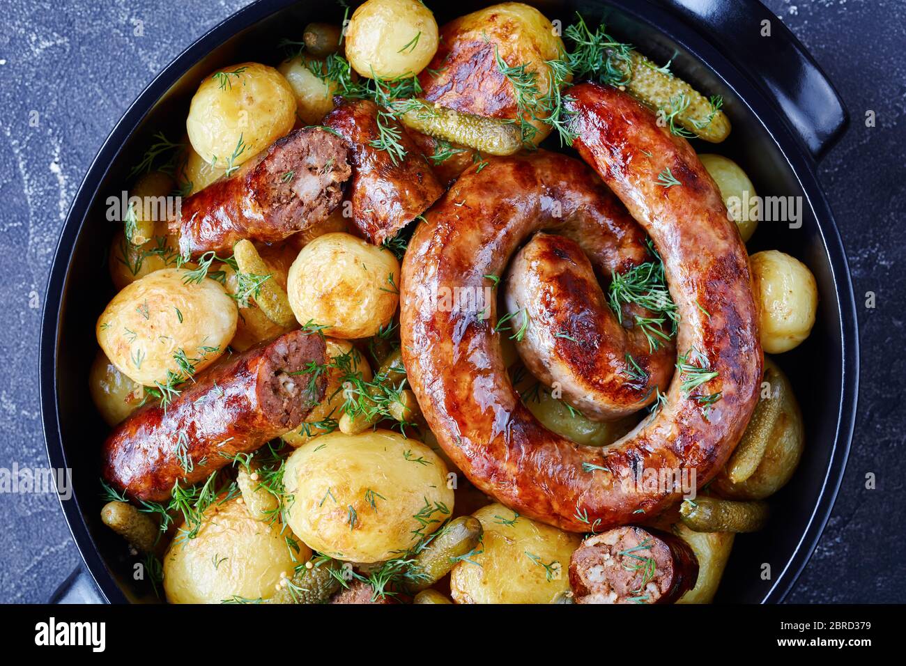 Roasted ring sausage and young potato with gherkins, sprinkled with fresh dill on a black dish on a dark concrete background, top view,  close-up Stock Photo