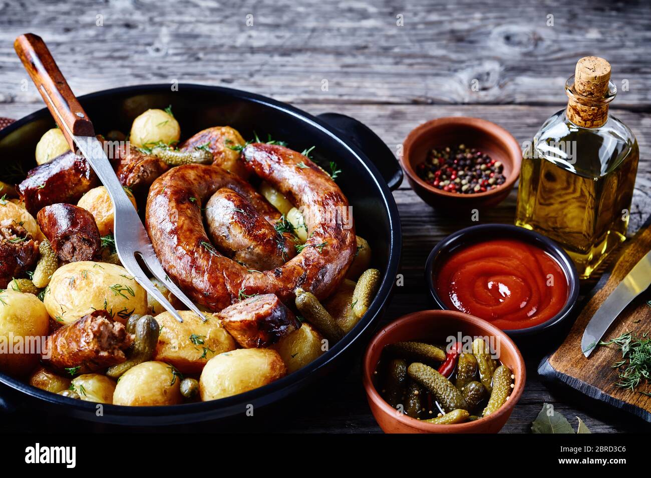 Easy pork sausage with young potato and gherkins, sprinkled with fresh dill on a black dish on a wooden background with ketchup, peppercorns, olive oi Stock Photo