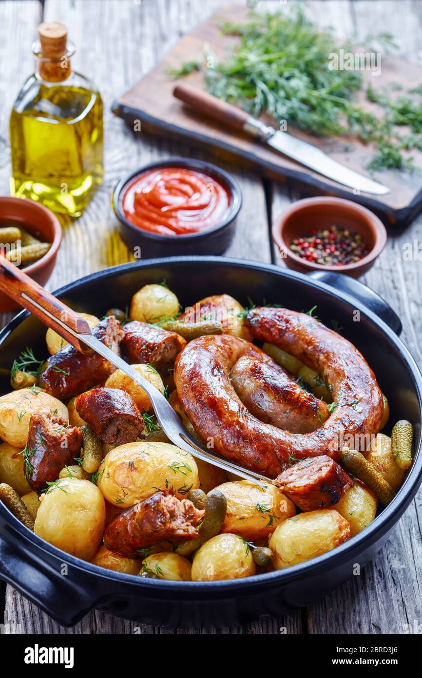 Oven-roasted spiral pork sausage with new potato and gherkins, sprinkled with fresh dill on a black dish on a wooden background with ketchup, pepperco Stock Photo