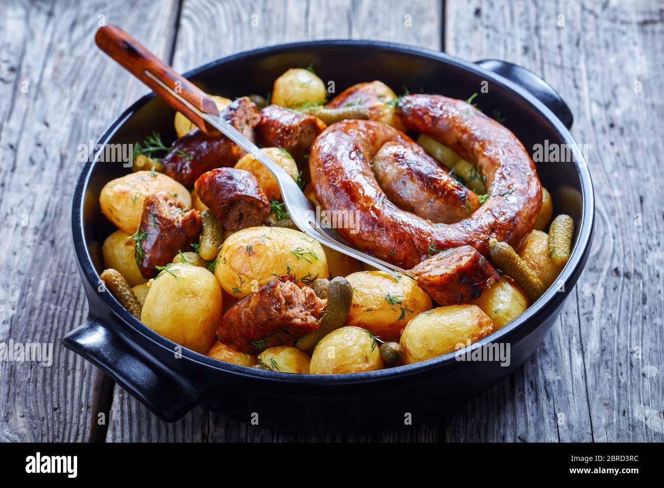 Italian sausage roasted with new potato and gherkins, sprinkled with fresh dill on a black baking dish on a wooden table, close-up Stock Photo