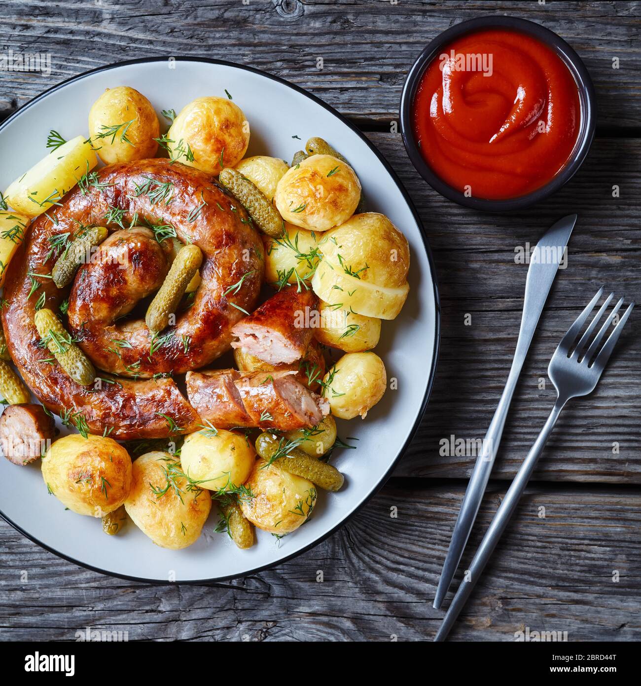 German lunch: new potato, sausage and gherkins, sprinkled with fresh dill on a plate with cutlery and ketchup on a wooden table, flat lay, close-up Stock Photo