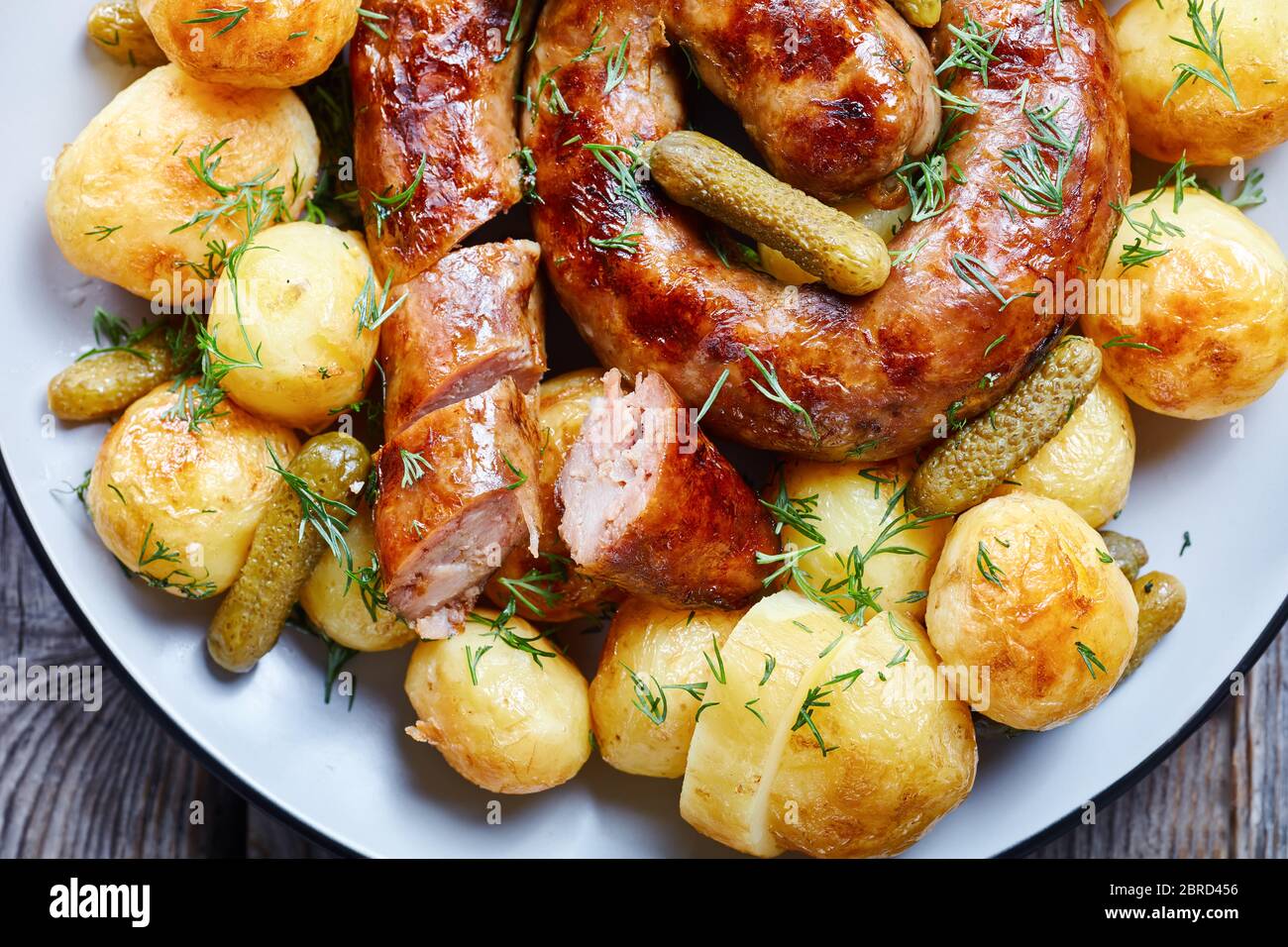 German food: new potato boiled and fried, served with round sausage and pickled cucumbers, sprinkled with fresh dill, served on a plate on an old wood Stock Photo