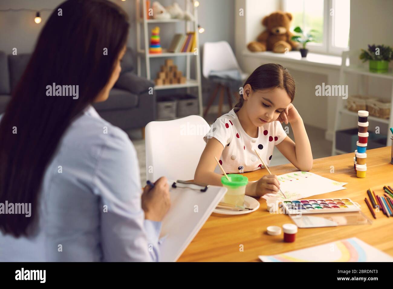 Child psychologist. A woman psychologist with a clipboard works with a little girl patient in a children's room. Psychological assistance analysis for Stock Photo