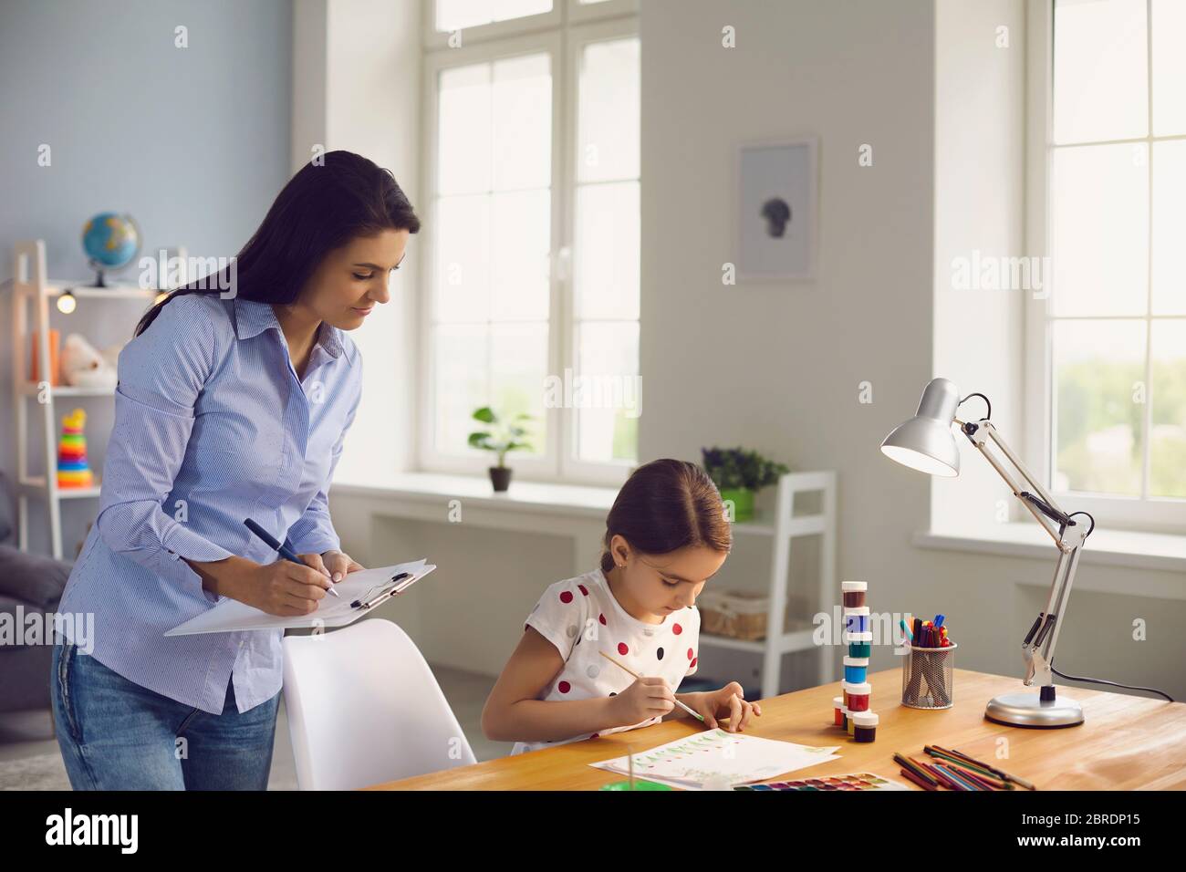 Child psychologist psychology. A woman psychologist with a clipboard works with a little girl patient in a children's room. The psychological assistan Stock Photo