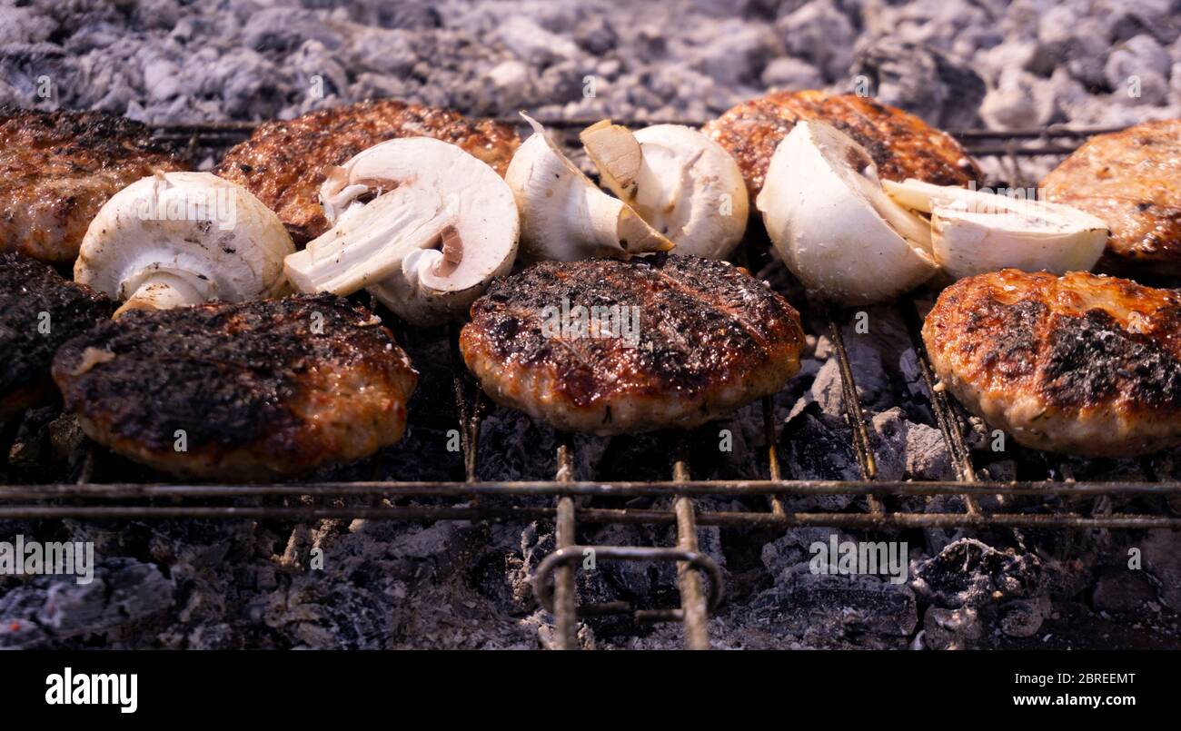 Beef BBQ Cooking of burger meat and mushroom. Iron barbeque grill pan with fire and food. Hamburgers outdoors barbecuing on coals. Closeup beef. Stock Photo
