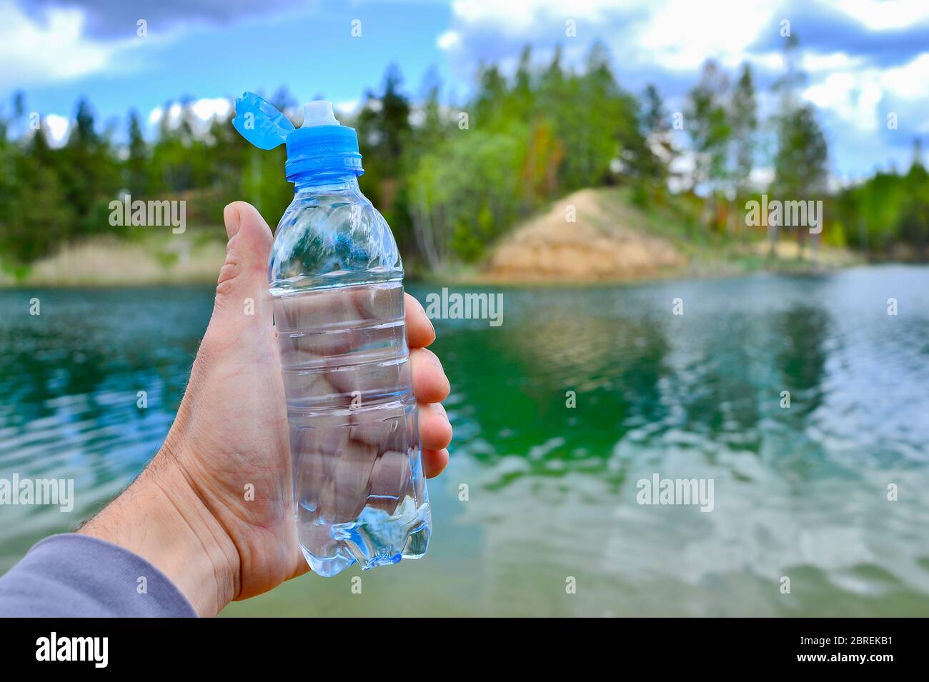 https://c8.alamy.com/comp/2BREKB1/a-mans-hand-holds-a-water-bottle-close-up-against-the-background-of-clear-water-of-the-lake-with-a-turquoise-hue-and-a-summer-landscape-background-2BREKB1.jpg