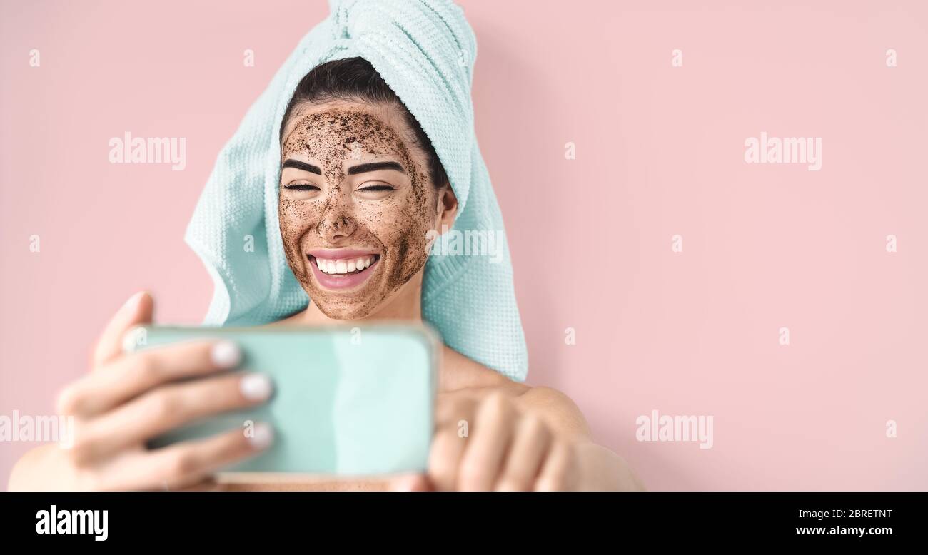 Young smiling woman taking selfie while doing coffee scrub facial mask - Happy girl having skin care spa day at home Stock Photo