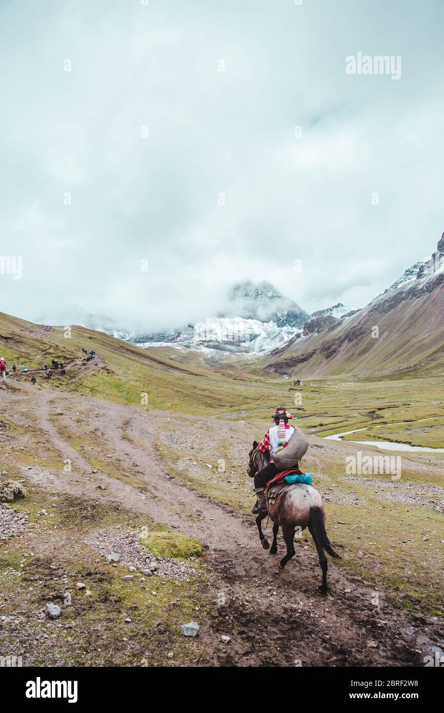 Traditional indigenous Quechua horseman rides his horse across the valleys of the Cusco region of Peru, next to Rainbow Mountain (or Montaña de Siete Stock Photo