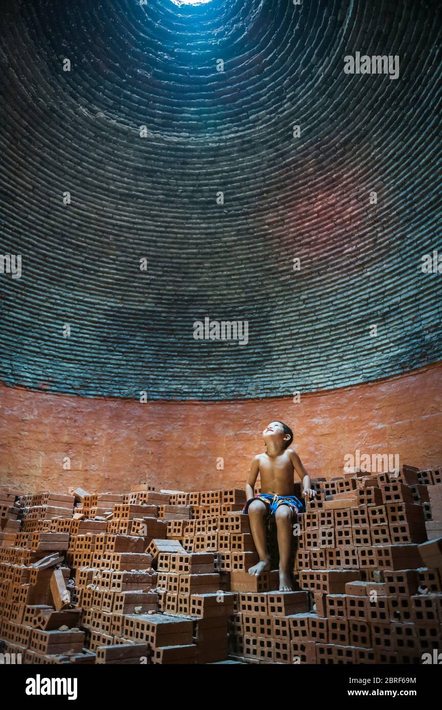 A young Cambodian boy sits on a pile of bricks looking up to the light coming through the hole in the roof of a beehive brick kiln. Battambang, Cambod Stock Photo