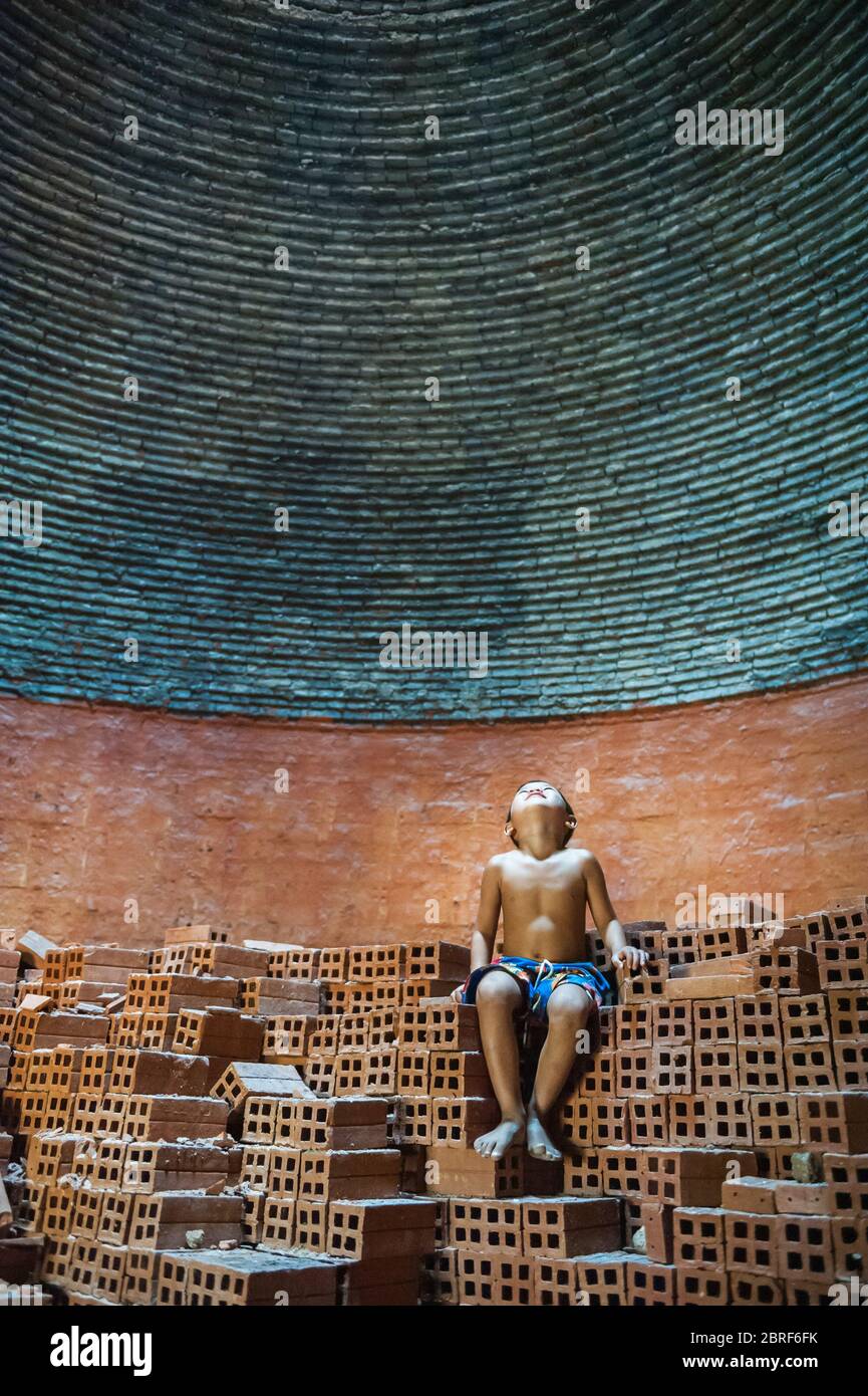 A young Cambodian boy sits on a pile of bricks looking up to the light coming through the hole in the roof of a beehive brick kiln. Battambang, Cambod Stock Photo