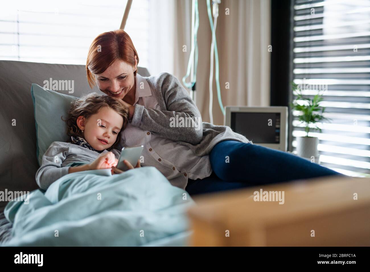 Small girl with mother in bed in hospital, using smartphone to pass time. Stock Photo