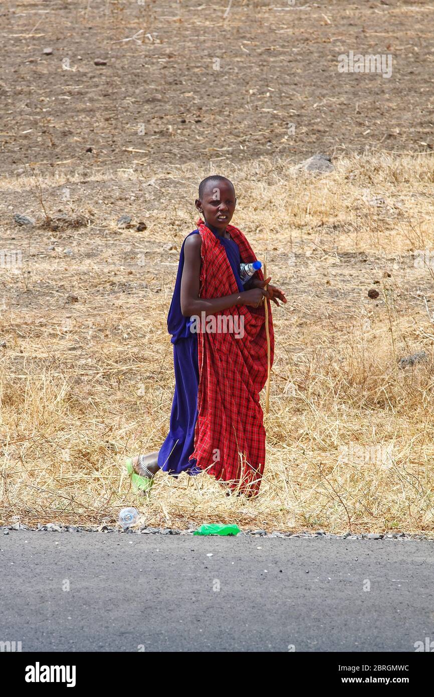https://c8.alamy.com/comp/2BRGMWC/maasai-boy-walking-by-road-carrying-water-bottle-stick-red-and-blue-shukas-traditional-dress-tribe-moving-tanzania-africa-2BRGMWC.jpg