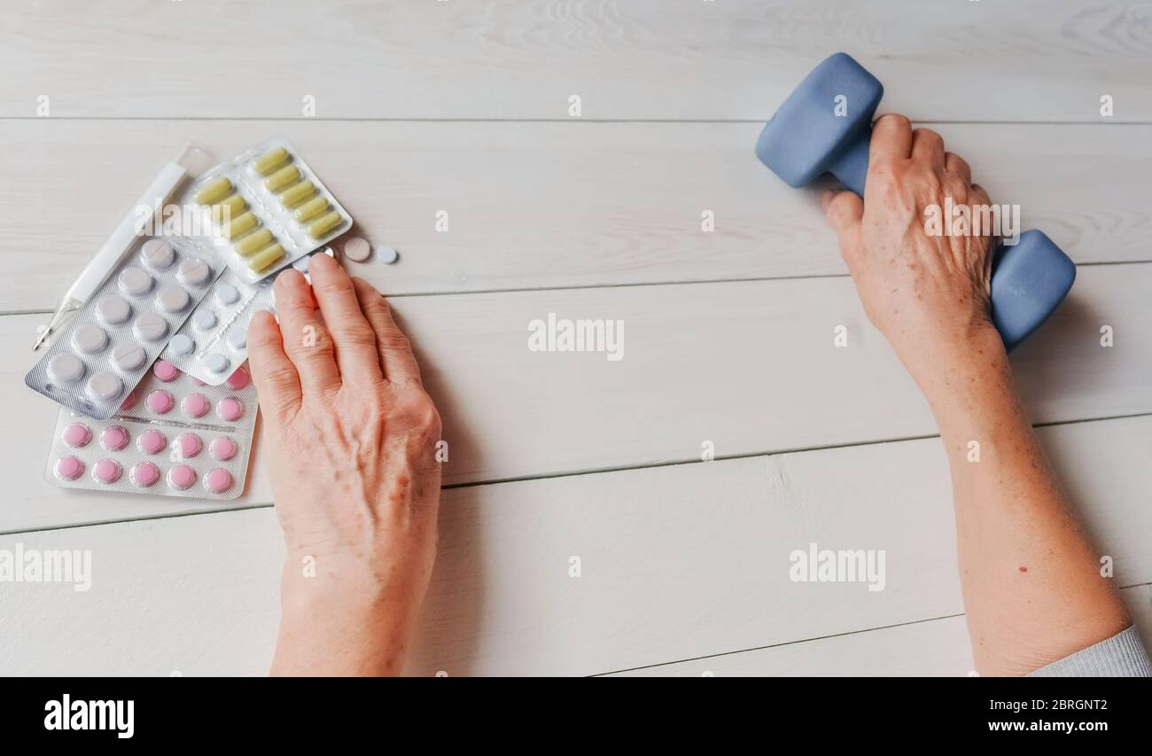 Senior hands with pills, drugs on table, dumb-bell. Woman wrinkled hands, thermometer, colorful tablets and dumb-bell, wooden background. Health care Stock Photo