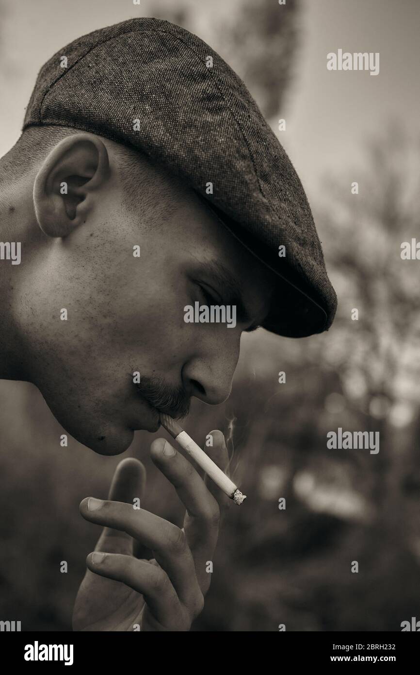 Retro 1920s portrait of an English gangster with a flat cap. Smokes a cigarette on the street.. Stock Photo