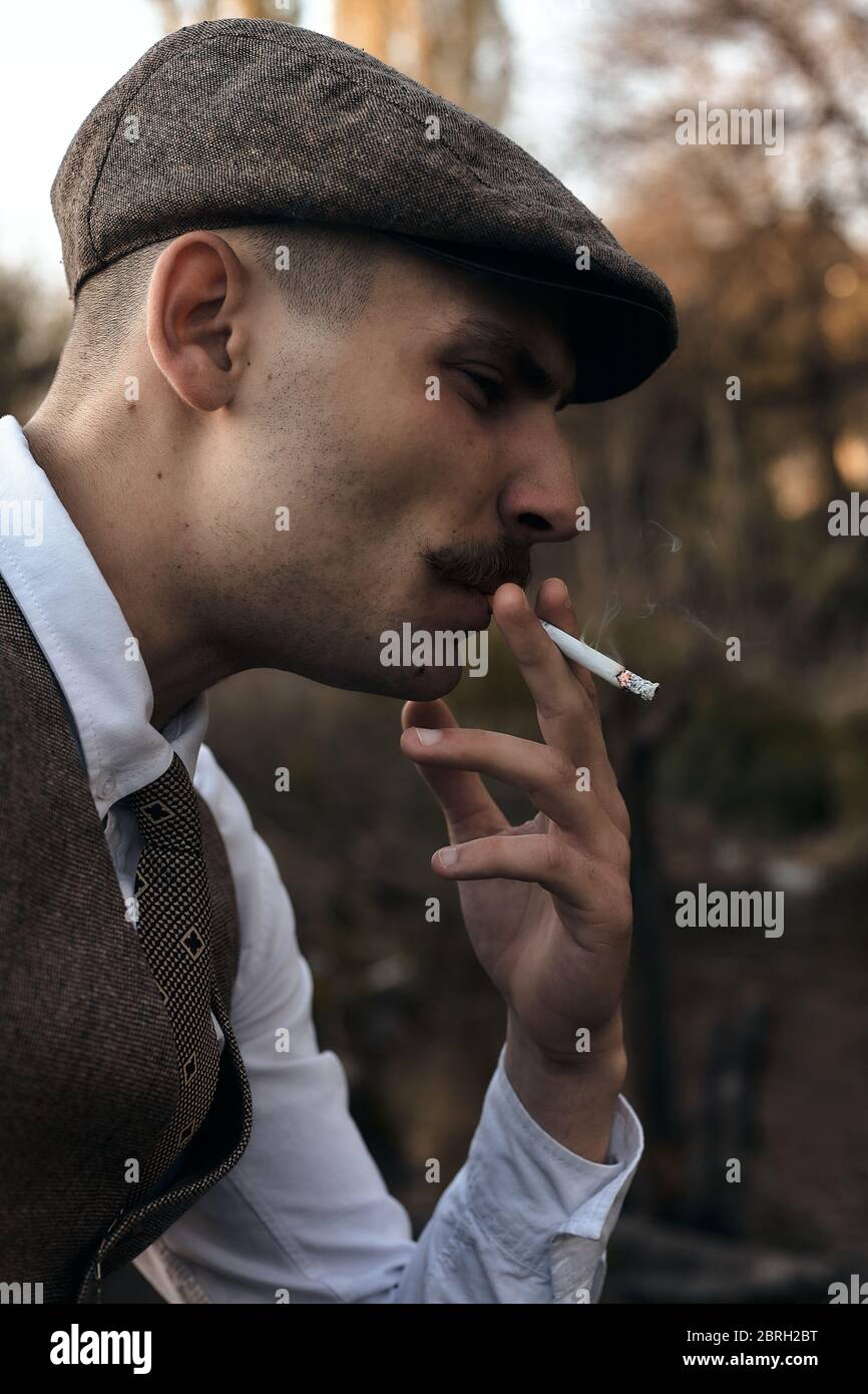 Retro 1920s portrait of an English gangster with a flat cap. Smokes a  cigarette on the street Stock Photo