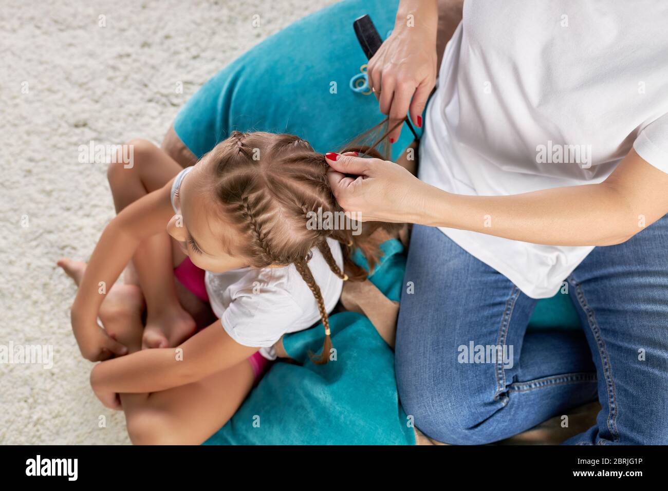 Mom makes hairdo her little daughter sitting on a soft chair, top view. Stock Photo