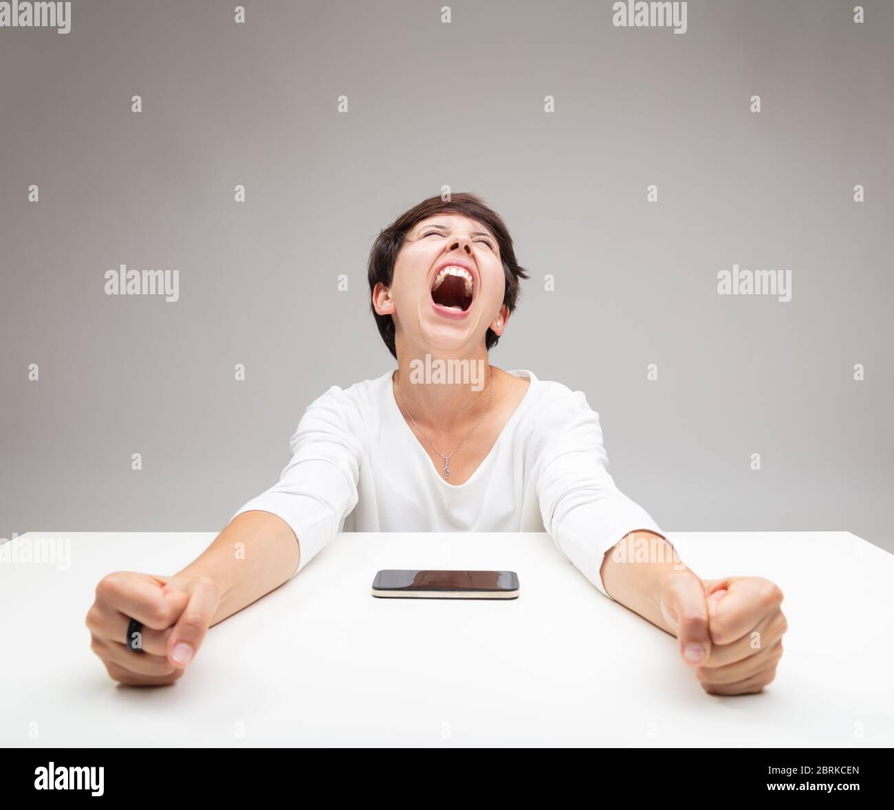 Frustrated woman throwing a temper tantrum clenching her fists and screaming at a mobile phone on the table in front of her Stock Photo