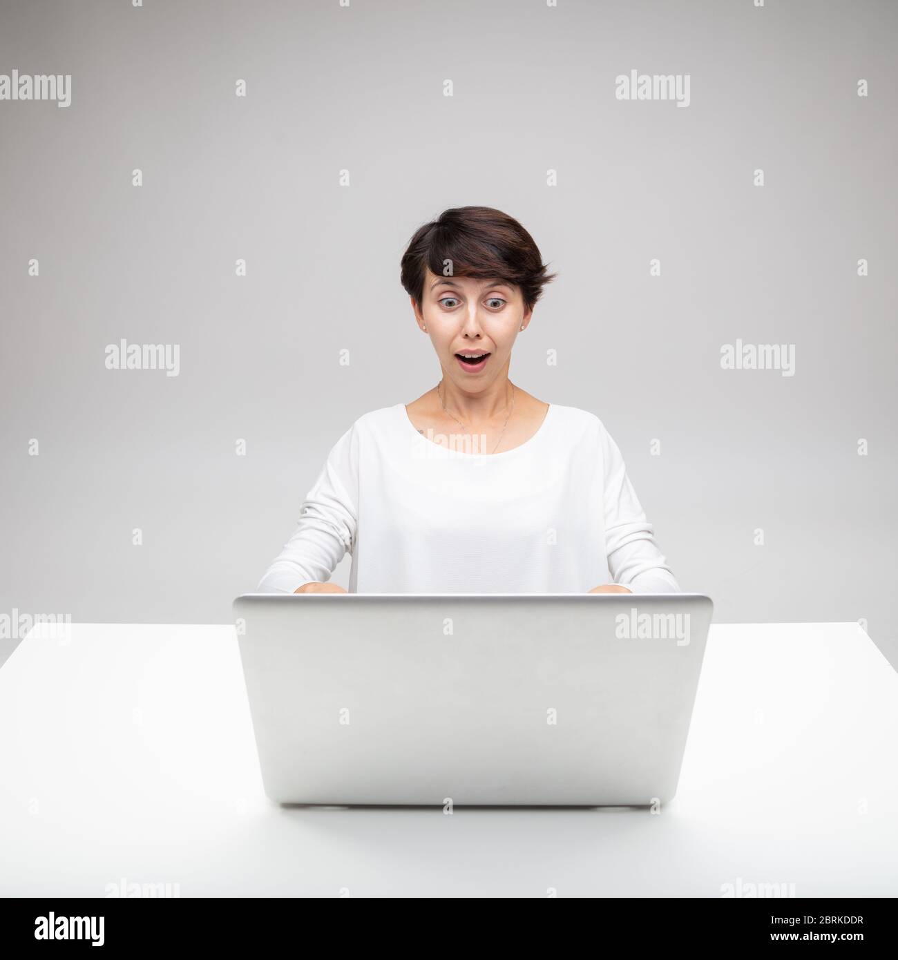 Astonished woman gawping at a laptop in amazement with wide eyes and mouth agape as she sits at a white desk over a grey studio background Stock Photo